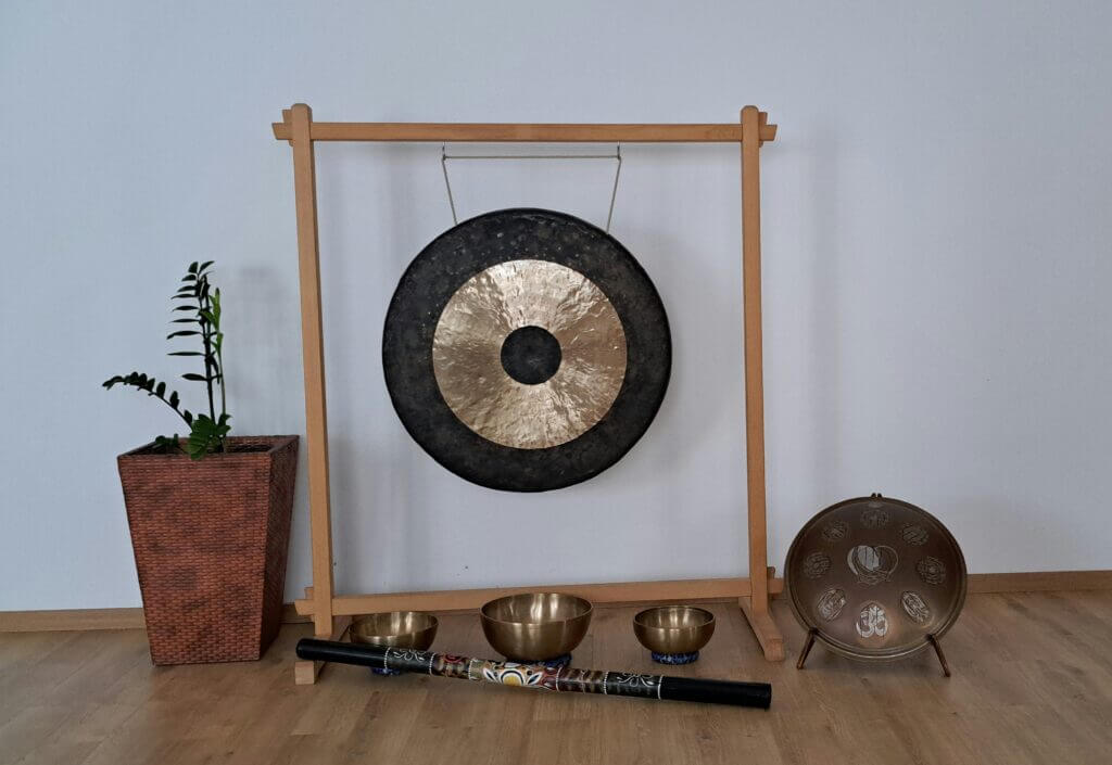 Hangutazás eszközei: gong, hangtálak és hangterápiás hangszerek egy meditatív térben Hangutazáshoz használt hangszerek, köztük egy nagy gong, három hangtál, egy hangcső és egy különleges kézidob, egy letisztult jógastúdió falánál elhelyezve